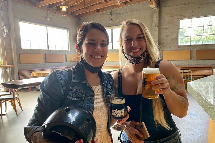 Two women standing next to each other and smiling while holding beer, hard cider, and a bike helmet