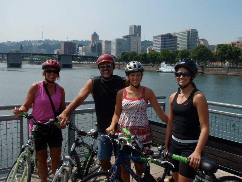 Group of 4 cyclists with bikes standjng together and smiling at the Portland waterfront with a downtown city view behind them