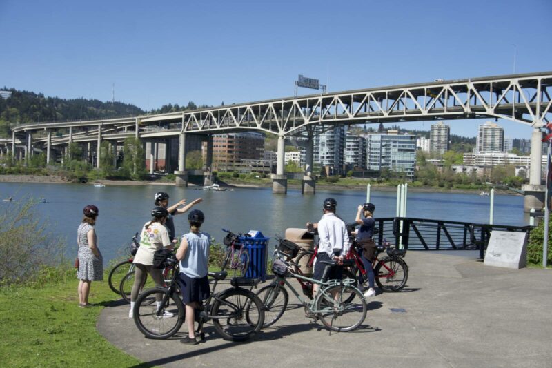 Group of cyclists stopped at the portland waterfront under a bridge while listening to their tour guide